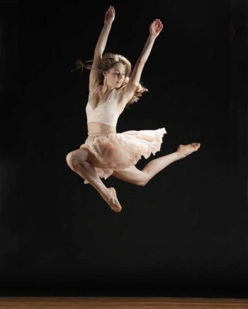 Dancer in a pale pink tutu leaping with arms raised against a black backdrop