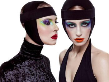 Two models in black headwraps with bold rainbow eye makeup and orange lips
