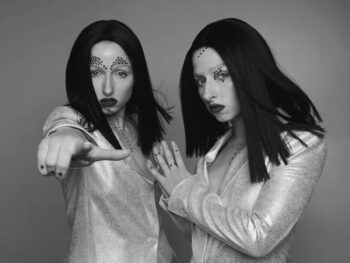 Two models with face gems in metallic jackets, one pointing at the camera, black-and-white