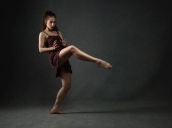 Dancer in a dark red dress balancing in a high kick against a dark backdrop