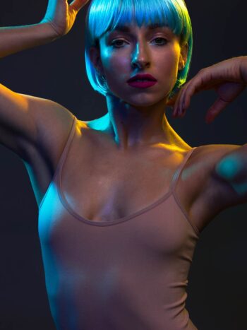 Model in a blue bob wig posing with raised arms under colored gel lighting