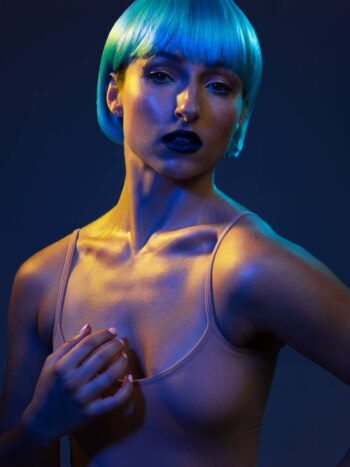 Model in a blue bob wig and nude bodysuit under colored gel lighting
