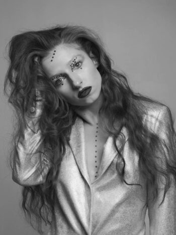 Model with long wavy hair and face gems in a metallic jacket, black-and-white